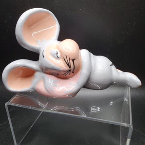 May include: A ceramic figurine of a gray and pink mouse sleeping on its side. The mouse has large pink ears and a closed eye.