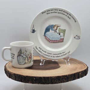 May include: A white ceramic teacup and saucer set featuring a Beatrix Potter illustration of Peter Rabbit in bed with his mother. The text on the saucer reads "Peter was not very well during the evening. His mother put him to bed." and the text on the teacup reads "and made some camomile tea: One tablespoonful to be taken at bedtime."