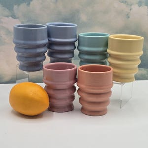 Puede incluir: Colección de seis jarrones de cerámica en colores pastel, incluyendo azul, rosa y amarillo. Cada jarrón tiene un diseño acanalado y una forma cilíndrica. Una fruta cítrica naranja se encuentra cerca de los jarrones.