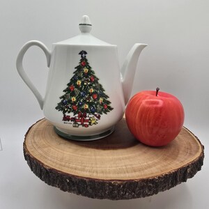 May include: White ceramic teapot with a Christmas tree design. The teapot has a green and white trim and a green lid. The tree is decorated with red and green ornaments, a red bow, and a star on top. The teapot is sitting on a wooden slice.