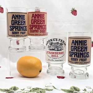 Może przedstawiać: Cztery przezroczyste, zabytkowe szklanki z etykietami. Dwie szklanki mają etykiety "Annie Green Springs Berry Frost", jedna "Annie Green Springs Country Cherry", a jedna "Boone's Farm Strawberry Hill". Pomarańcza leży z przodu.