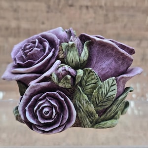 Puede incluir: Una escultura detallada de rosas y capullos morados con hojas verdes. El arreglo floral está intrincadamente diseñado, mostrando la textura de los pétalos y las hojas. La escultura se encuentra sobre un fondo borroso.