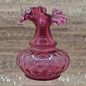 Puede incluir: Un jarrón de cristal color arándano vintage con un borde con volantes. El jarrón tiene un cuerpo bulboso con una superficie texturizada y estampada. El borde con volantes añade un toque delicado al diseño antiguo. El jarrón es una pieza decorativa.