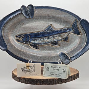 May include: A blue and white ceramic oval serving dish with a raised fish design. The dish is on a wooden stand with two business cards. The business cards read "Blue Spruce Pottery" and "Studio & Gallery".