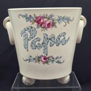 May include: Cream ceramic planter with the word "Papa" in blue floral lettering. It features pink roses and blue floral accents, small handles, and three ball feet. A decorative item for the home, ideal for flowers or plants.