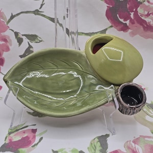 Puede incluir: Una fuente de cerámica verde en forma de hoja con una sección en forma de oliva y una pequeña taza marrón. La fuente está sostenida por soportes de plástico transparente. El fondo es un estampado floral.
