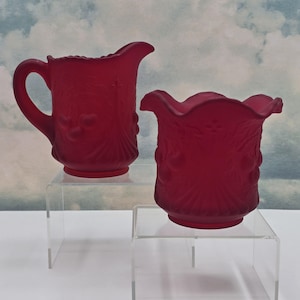 May include: Two pieces of ruby red glass tableware. A small jug with a handle and a ruffled rim, and a matching open-top container. Both have a frosted finish and a raised floral design.