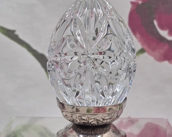 May include: A clear, faceted glass egg ornament with a detailed floral pattern. The egg sits on a silver-colored metal base with an ornate design. The background features a floral pattern.