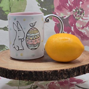 May include: White ceramic Easter mug with a bunny and egg design, a pink interior, and a decorative handle. A whole yellow lemon sits next to the mug on a round wooden serving board.