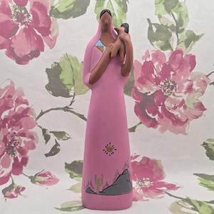May include: A pink ceramic figurine of a woman holding a child. The woman wears a long dress with a desert landscape design. The design includes a sun, mountains, and a cactus. The figurine has a minimalist style.