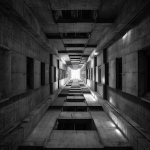 Puede incluir: Fotografía en blanco y negro de una estructura de hormigón, vista desde abajo, creando una perspectiva de profundidad. La imagen presenta un largo espacio rectangular en forma de túnel con patrones geométricos repetidos y una luz brillante al final.