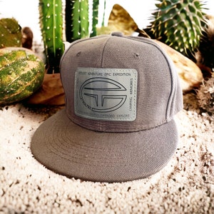 May include: A gray baseball cap with a silver patch that says "GETLOST ADVENTURE EPIC EXPEDITION" and "TSOLTEC" with a logo of a circle with a T inside. The patch also says "CAMPING MEMORIES" and "OVERLAND FAMILY OFFROAD EXPLORE".