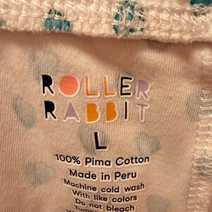 Puede incluir: Primer plano de una tela blanca con un patr&oacute;n repetido de corazones turquesa. El texto "ROLLER RABBIT" est&aacute; impreso en letras coloridas. Debajo, la etiqueta dice "L" y "100% Pima Cotton Made in Peru". Tambi&eacute;n se imprimen instrucciones de lavado.