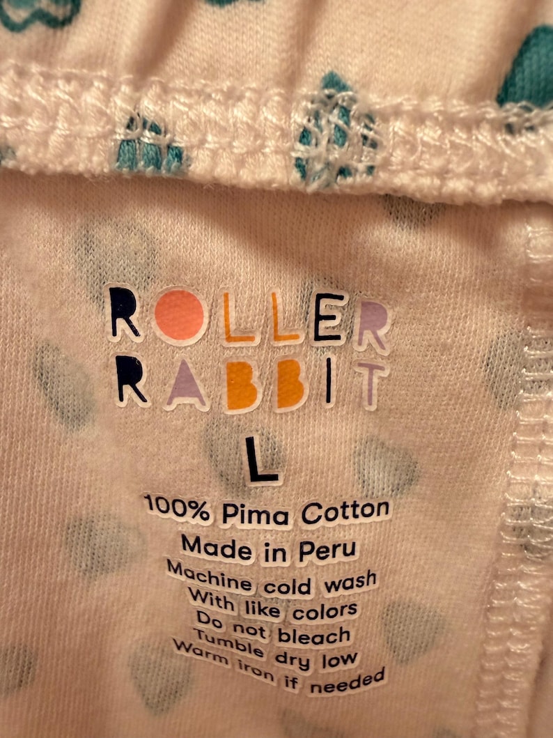 Puede incluir: Primer plano de una tela blanca con un patr&oacute;n repetido de corazones turquesa. El texto "ROLLER RABBIT" est&aacute; impreso en letras coloridas. Debajo, la etiqueta dice "L" y "100% Pima Cotton Made in Peru". Tambi&eacute;n se imprimen instrucciones de lavado.