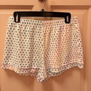 Puede incluir: Pantalones cortos de pijama blancos con un estampado de corazones en verde azulado, rojo y morado. Los pantalones cortos tienen un ribete morado y cuelgan de una percha negra.