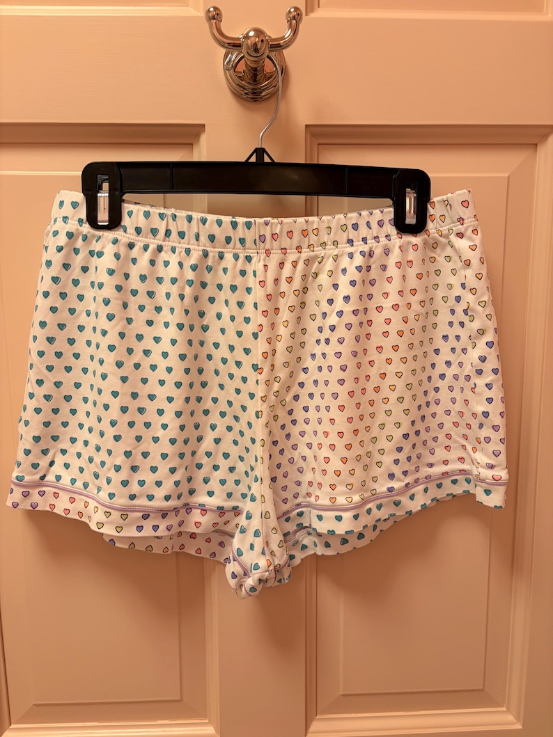 Puede incluir: Pantalones cortos de pijama blancos con un estampado de corazones en verde azulado, rojo y morado. Los pantalones cortos tienen un ribete morado y cuelgan de una percha negra.