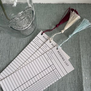 May include: A set of three white reading list bookmarks with black text and lines. The bookmarks have a tassel at the top in burgundy, cream, and light blue.