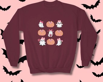 Puede incluir: Sudadera burdeos con un diseño de fantasmas y calabazas de dibujos animados con lazos rosas y detalles florales. El texto "Available in other colors!" está en la parte inferior, con opciones de color. Siluetas de murciélagos negros sobre fondo rosa.