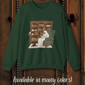 May include: Dark green sweatshirt featuring a cartoon ghost reading in a green armchair in front of a bookshelf. A black cat is lying on the rug. The text "Available in many colors!" is at the bottom, with color options below.