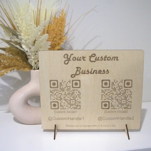 May include: A wooden sign with the text "Your Custom Business" and two QR codes. The text below the QR codes reads "Custom Social1 @CustomHandle1" and "Custom Social2 @CustomHandle2". The sign is on a small wooden stand.