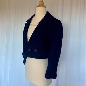 May include: A cropped navy blue blazer with a double breasted closure and long sleeves. The blazer has a pearl necklace draped over the collar.