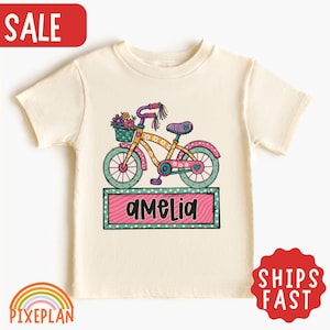 May include: A cream-colored short-sleeve t-shirt featuring a graphic of a colorful bicycle with a flower-filled basket on the handlebars. Below the bicycle is a rectangular pink and green frame containing the name Amelia in black script.