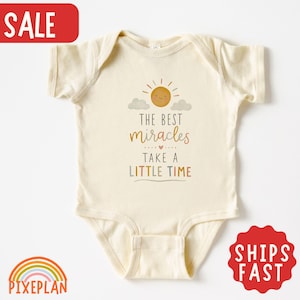Könnte beinhalten: Ein cremefarbener Kurzarm-Babybody mit einer Grafik aus einer lächelnden Sonne und Wolken. Der Text lautet: THE BEST miracles TAKE A LITTLE TIME. Das Design verwendet gedeckte Töne in Orange, Braun und Blau für die Schrift und die Sonnenillustration.
