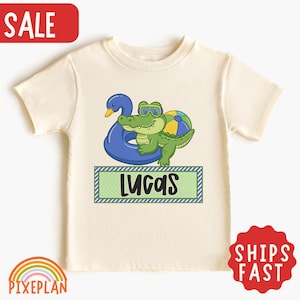 May include: A cream-colored short-sleeve t-shirt featuring a graphic of a green crocodile wearing goggles, resting on a blue swan-shaped pool float next to a beach ball. Below the graphic is a green rectangular box with the name LUCAS in black.