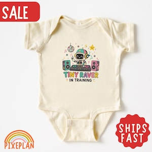 May include: A cream-colored short-sleeve baby bodysuit featuring a colorful graphic of a robot DJ with headphones, speakers, a disco ball, and stars. The text below the graphic reads "TINY RAVER IN TRAINING" in multi-colored block letters.