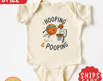 Peut inclure: Un body pour bébé à manches courtes de couleur crème avec un motif de ballon de basket animé portant un bandeau, en train de marquer un panier. Le texte HOOPING & POOPING est inscrit en lettres bleu foncé et orange sur la poitrine.