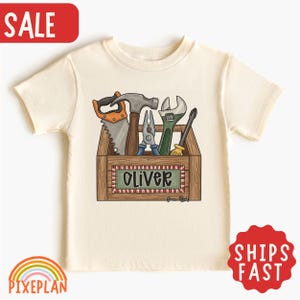 May include: A cream-colored short-sleeve t-shirt featuring a graphic of a wooden toolbox filled with various hand tools including a saw, hammer, pliers, wrench, and screwdriver. The front of the toolbox displays a green label with the name Oliver.