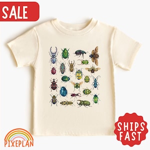 Puede incluir: Una camiseta de manga corta de color crema con un estampado gráfico central de varios escarabajos e insectos coloridos en tonos verdes, azules, marrones y amarillos. El diseño muestra una colección de diferentes especies en un patrón.