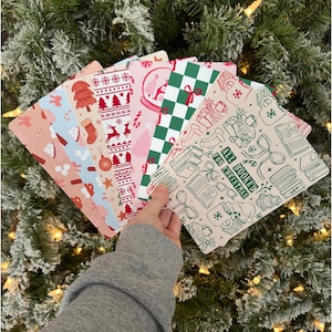 May include: A set of rectangular bookmarks featuring various holiday-themed patterns including red Christmas trees, stockings, green checkered prints, and a cream-colored card with the text ALL BOOKED FOR CHRISTMAS surrounded by illustrations of books and mugs.