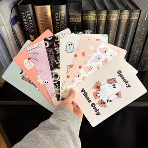 May include: A set of eight Halloween-themed decorative cards featuring a variety of ghost illustrations. The cards are pink, blue, and white with black and orange accents. The text on one card reads "Spooky Vibes Only".