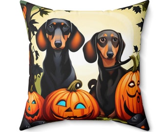 May include: A square throw pillow featuring a graphic illustration of two black and tan dachshund dogs sitting among three carved orange jack-o'-lantern pumpkins with glowing blue eyes and mouths against a pale yellow background with dark leaves.