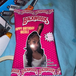 May include: A pink Backwoods leaf wrapper with the text "HAPPY BIRTHDAY NICOLE" and a photo of a woman. The wrapper also says "HONEY BERRY CIGAR" and "ALL NATURAL LEAF WRAPPER". Hershey's chocolate bars and scissors are in the background.