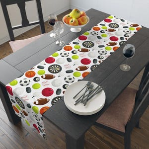 May include: A white table runner featuring a repeating pattern of various sports equipment including basketballs, footballs, soccer balls, baseballs, bowling balls, dartboards, hockey pucks, tennis balls, and volleyballs printed across the fabric surface.