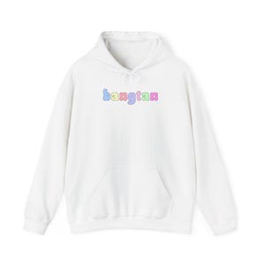 Könnte beinhalten: Ein weißer Kapuzenpullover mit einer Vordertasche und einem Kordelzug. Das Wort "bangtan" ist in Pastellfarben, darunter Blau, Gelb und Lila, auf die Brust gedruckt. Der Pullover ist aus weichem Material.