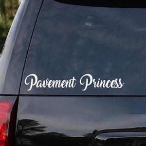 May include: A white vinyl decal applied to the rear window of a black vehicle. The decal features the text Pavement Princess written in a cursive script font. The lettering is positioned horizontally across the lower section of the glass.