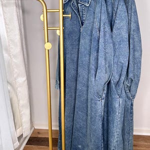 May include: Three denim dresses hanging on a gold metal clothing rack with a white marble base. The dresses are all the same style and color, with a slightly distressed look.