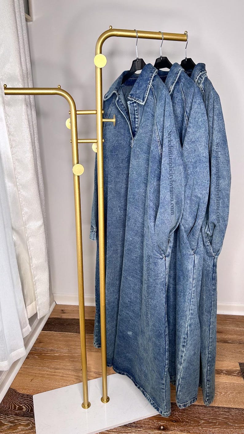 May include: Three denim dresses hanging on a gold metal clothing rack with a white marble base. The dresses are all the same style and color, with a slightly distressed look.