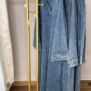 May include: A gold metal clothing rack with a white marble base. Three denim dresses hang on the rack.