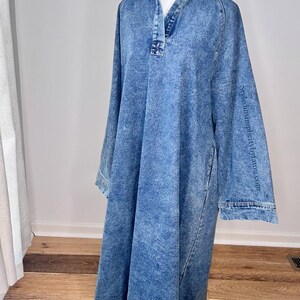 May include: A long-sleeved denim dress with a collared neckline and a button closure. The dress has a relaxed fit and a slightly A-line silhouette. The fabric has a light blue wash and a slightly distressed look. The dress is shown on a mannequin against a white background.
