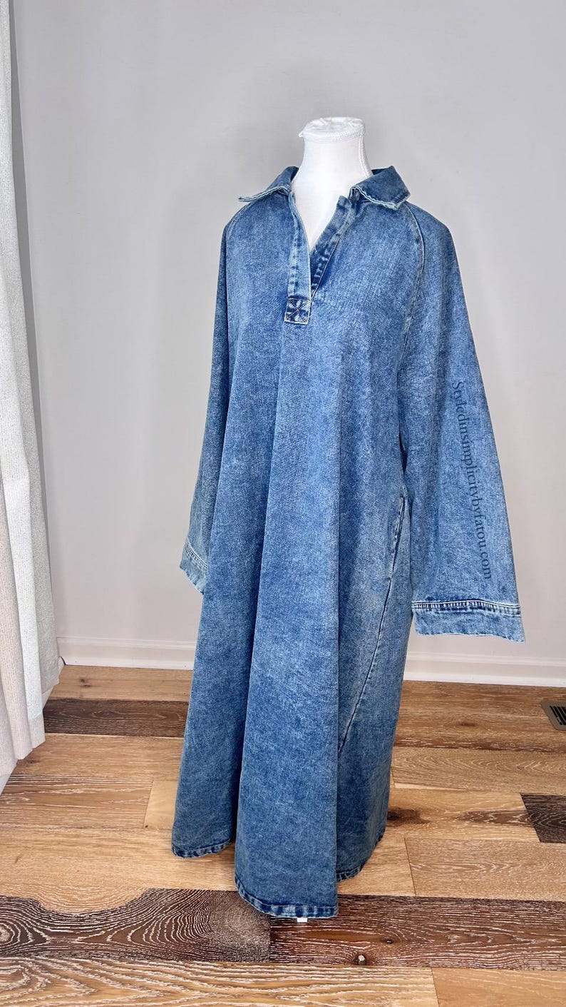 May include: A long-sleeved denim dress with a collared neckline and a button closure. The dress has a relaxed fit and a slightly A-line silhouette. The fabric has a light blue wash and a slightly distressed look. The dress is shown on a mannequin against a white background.