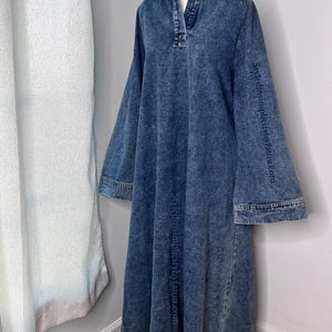May include: A long-sleeved denim dress with a collared neckline and a relaxed fit. The dress has a faded blue color and a slightly distressed look. The dress is on a white mannequin and the text "styledsimplybytatou.com" is printed on the sleeves.