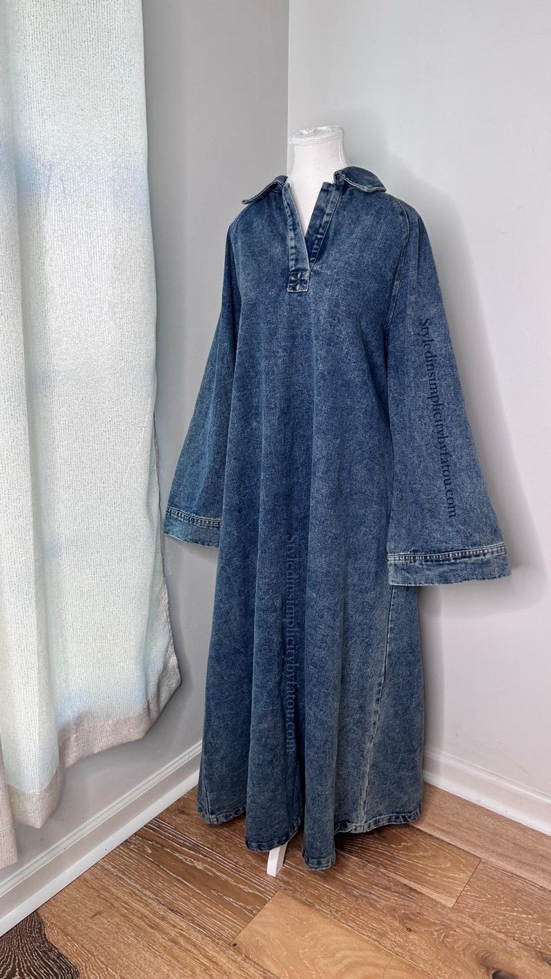 May include: A long-sleeved denim dress with a collared neckline and a relaxed fit. The dress has a faded blue color and a slightly distressed look. The dress is on a white mannequin and the text "styledsimplybytatou.com" is printed on the sleeves.