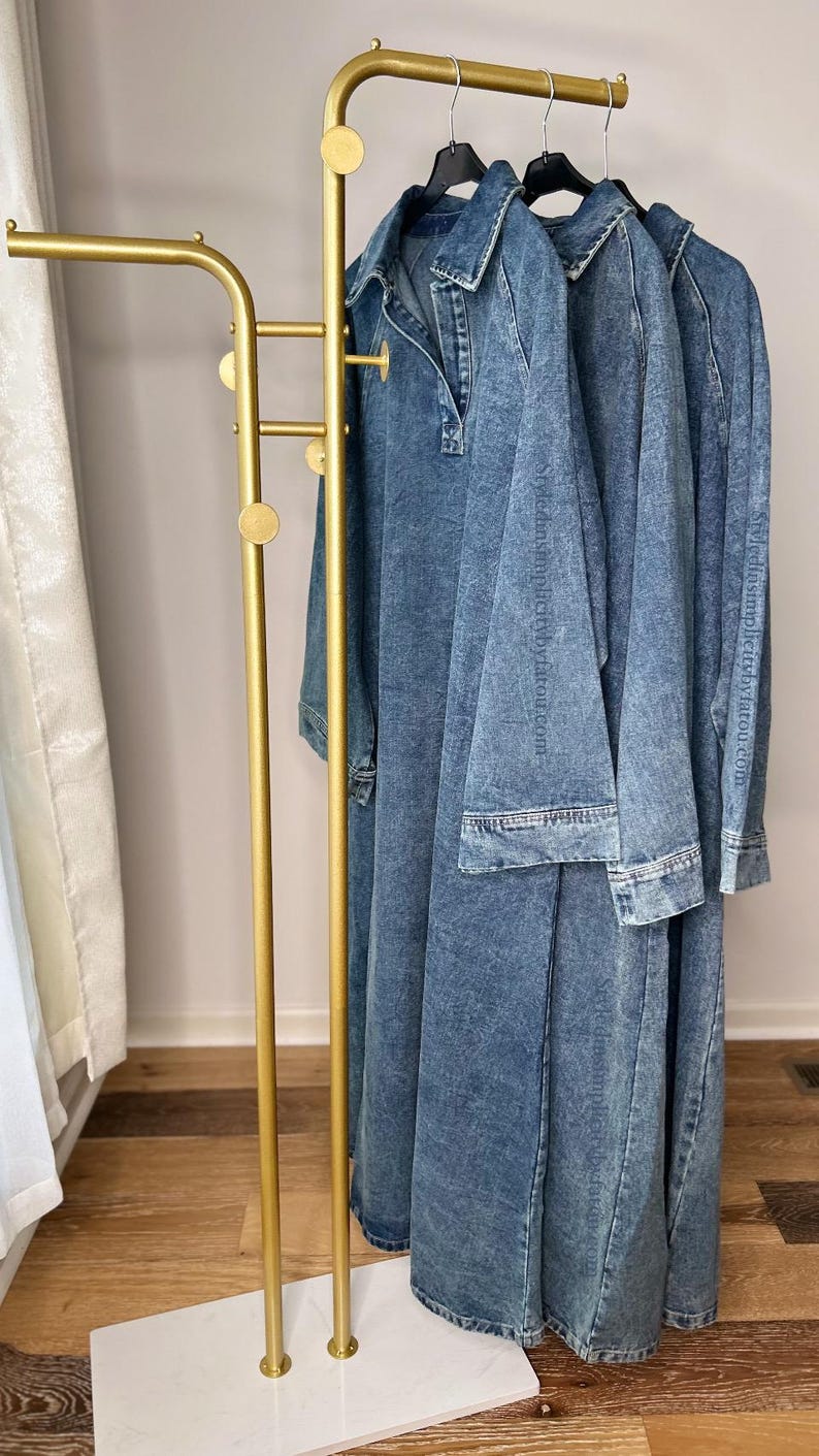 May include: A gold metal clothing rack with a white marble base. Three denim dresses hang on the rack.