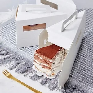 Puede incluir: Una porción de pastel en capas con cobertura de cacao en polvo se presenta en una caja triangular blanca con asa. El pastel es parcialmente visible, mostrando sus múltiples capas. Un tenedor dorado descansa sobre una superficie gris con flecos.