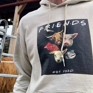 Op de afbeelding: Beige hoodie met een afbeelding met het woord "FRIENDS" boven afbeeldingen van een koe, varken, kip, lam en vis. De tekst "NOT FOOD" staat eronder gedrukt. De hoodie heeft een capuchon met trekkoord en een voorzak.