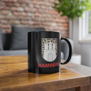 Puede incluir: Taza de café de cerámica negra con un diseño de estampado de leopardo blanco y marrón que presenta el escudo de armas de la ciudad de Hambourg. El texto "HAMBURG" está impreso en rojo en la taza.
